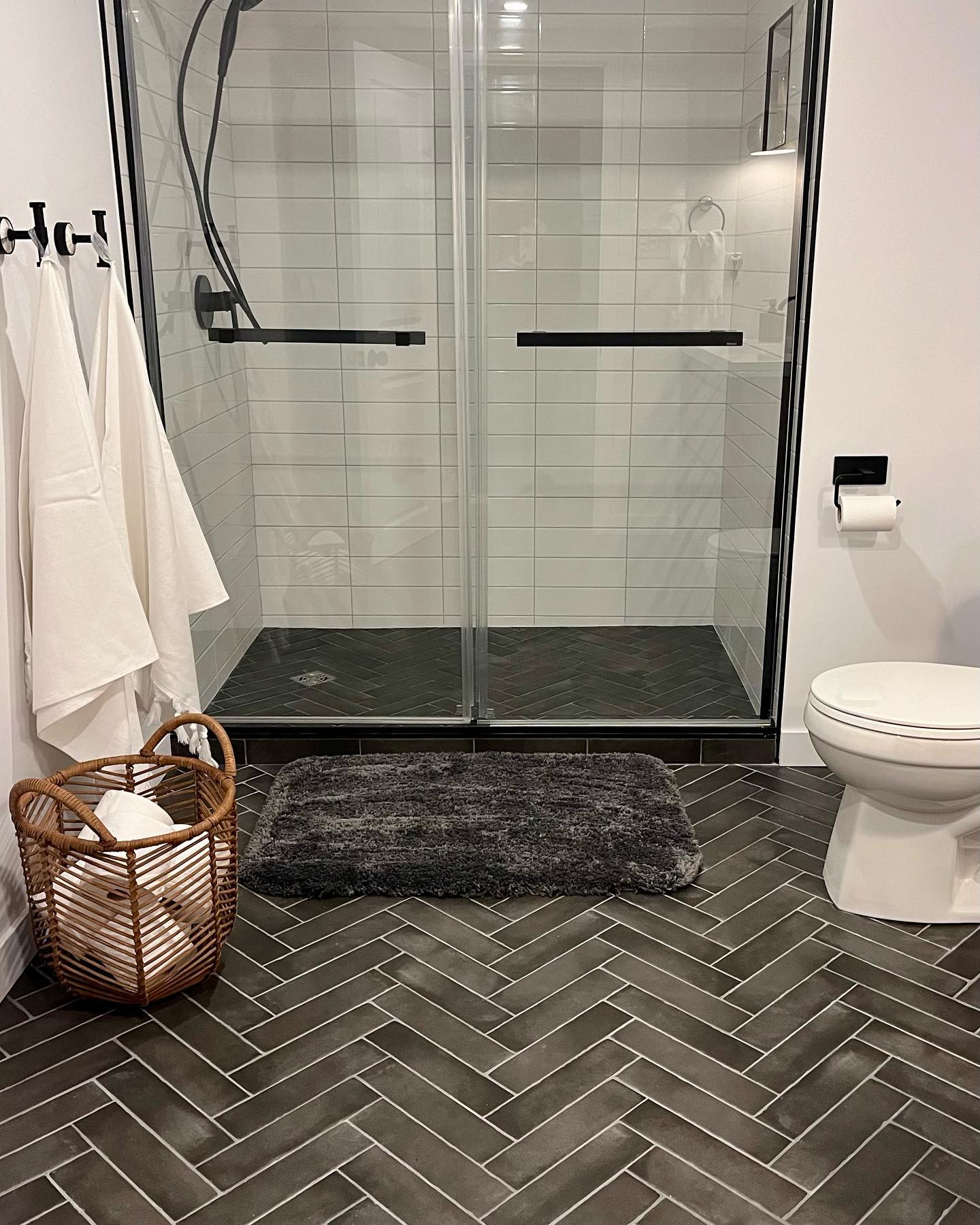 New basement bathroom with dark gray herringbone floor tile, black framed glass shower, and white subway tile walls