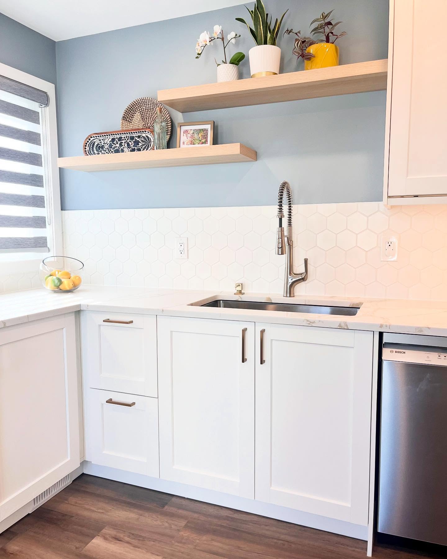 Finished Project Daisy sink wall with white shaker cabinets, hexagon tile, dusty blue paint, wood shelves, and a coil faucet