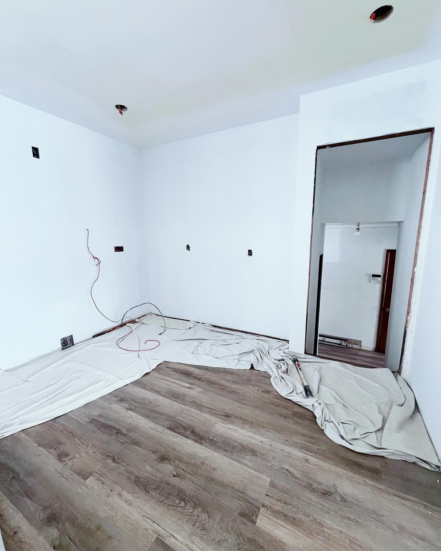 Project Rose room in progress with fresh white paint, new plank flooring, drop cloths, and open electrical boxes